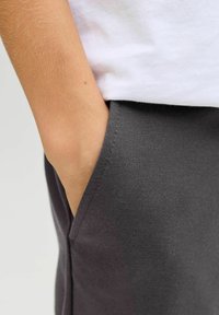 Main partiellement glissée dans la poche avant d'un pantalon gris foncé, portant une chemise blanche.