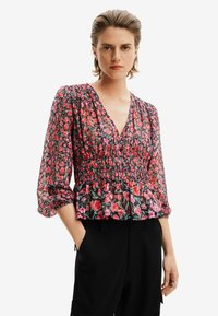 Blusa peplum floreale in tessuto nero trasparente con fiori rossi e rosa, caratterizzata da scollo a V e maniche a sbuffo. La texture sembra leggera.
