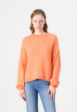Pull en tricot orange, duveteux, avec un col rond et des épaules tombantes, présentant des manches allongées et une coupe décontractée. Associé à un jean bleu.