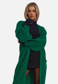 Cardigan verde a maglia con maniche oversize, tasche frontali e design aperto, indossato sopra un top e una gonna neri.