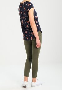 Námořnická květinová blůza s krátkými rukávy, spárovaná s úzkými olivově zelenými kalhotami a bílými teniskami. Blůza má zakřivený spodní lem a boční rozparky.