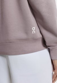 Sudadera morada suave con textura acanalada, con un logo blanco en la parte inferior. Combinada con pantalones blancos, resaltando un look casual.