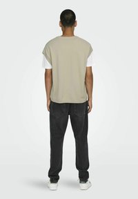 Beige ärmelloses Oberteil über einem weißen T-Shirt, kombiniert mit schwarzen Jeans. Das Oberteil hat eine lockere Passform und gerippte Akzente an den Schultern.