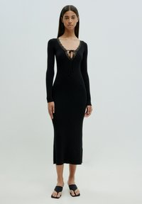 Zwarte geribbelde midi-jurk met lange mouwen, diepe V-hals met kantafwerking en strikdetail. Gepaard met zwarte slipper sandalen.