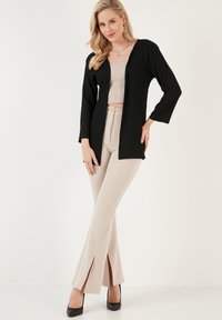 Cardigan noir à côtes avec manches longues, design à l'avant ouvert. Pantalons beiges taille haute, silhouette évasée avec fentes latérales. Talons noirs.