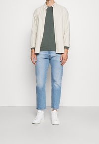 Camisa beige de botones sobre una camiseta verde oscura, jeans azul claro y zapatillas blancas. Conjunto casual con un ajuste relajado y texturas ligeras.