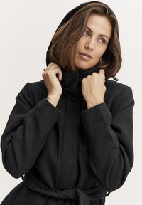 Cappotto nero con cappuccio e colletto alto, in tessuto текстurizzato e con vita legata. Presenta bottoni al collo e maniche lunghe e morbide.