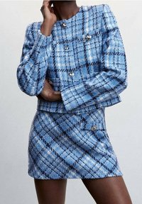 Nainen sinivalkoisessa ruudullisessa tweed-takissa hopeisilla napeilla ja siihen sopivassa minihameessa, poseeraa käsi kasvojen lähellä.