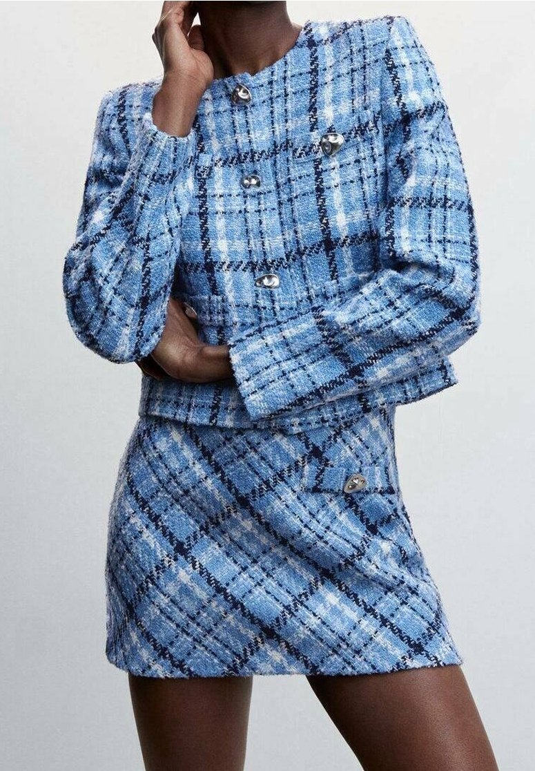 Nainen sinivalkoisessa ruudullisessa tweed-takissa hopeisilla napeilla ja siihen sopivassa minihameessa, poseeraa käsi kasvojen lähellä.