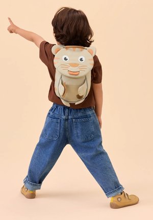 Child wearing a tiger-shaped backpack points forward, dressed in brown shirt, blue jeans, and tan shoes on a beige background.