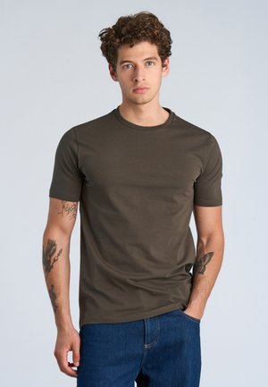 Ung mand med krøllet hår iført en tætsiddende brun t-shirt og blå jeans, stående med den ene hånd i lommen mod en ensfarvet baggrund.