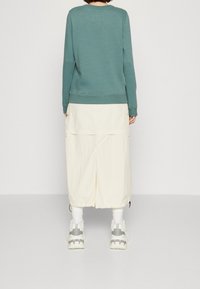 Felpa verde menta con polsini a coste, abbinata a una gonna beige chiaro al ginocchio con tasche laterali e orlo regolabile. Calzini bianchi e sneakers robuste.