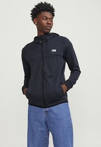 Tengerészkék cipzáros pulóver elülső zsebbel és húzózsinórokkal. Kontrasztos anyagpanel a mellrészen, apró logó a bal oldalon.