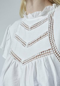 Hvid bluse lavet af letvægtsstof, med en rynket hals, indviklet blonde detaljer og flydende ærmer med en løs pasform.