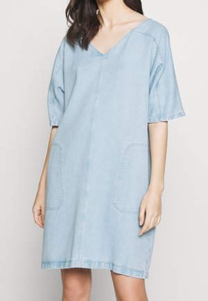 Žena v volných světle modrých džínových šatech s výstřihem do V, krátkými širokými rukávy a dvěma velkými předními kapsami, stojící před jednobarevným pozadím.