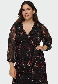 Mujer con cabello largo y oscuro vistiendo un vestido negro de mangas transparentes con patrones florales en rojo, marrón y blanco, y detalles de volantes, de pie con una mano en la cadera.