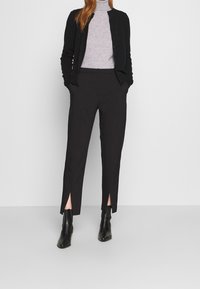 Kvinna iklädd svart cardigan, grå polotröja, svarta byxor med sprund vid anklarna och svarta ankelskor mot en enfärgad ljusgrå bakgrund.