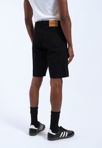Person iført sorte denimshorts med en brun label, sorte sokker, sort-hvide Adidas sneakers og en hvid ærmeløs skjorte.