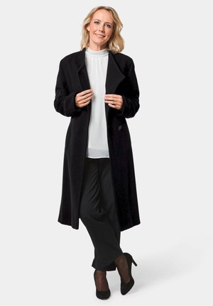 Long manteau noir en tissu épais et texturé, doté d'un col cranté et de poches latérales, superposé sur une blouse blanche et un pantalon noir.