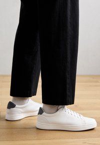 Baskets blanches en cuir avec des accents gris, des panneaux texturés et un design épuré. Associées à un pantalon noir court. Fond en parquet.