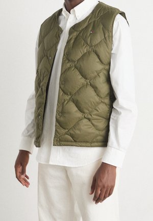 Man wearing an olive green quilted vest over a white button-up shirt and white pants, standing against a plain background.