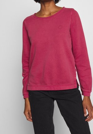 Personne portant une chemise à manches longues rose foncé délavée avec un col à bords bruts et un pantalon noir, sur un fond gris clair uni.