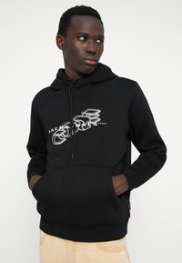 Svart hoodie med framficka, med en vit grafisk design och dragsnöre. Tillverkad av bomullsmaterial, med ribbade muddarn.