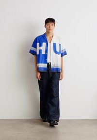 Lyhythihainen paita sinivalkoisena, jossa on suurta "HU"-kirjaimistoa, raidallisia yksityiskohtia, yhdistettynä pimeisiin leveisiin farkkuihin ja mustiin kenkiin.