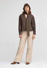 Blusa marrone con dettaglio a cravatta, aderente alle spalle e con maniche ampie; abbinata a pantaloni beige a gamba larga e stivaletti con tacco marroni.