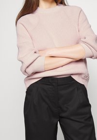 Femme aux longs cheveux portant un pull tricoté rose clair et un pantalon noir, les bras croisés à la taille, sur un fond uni.