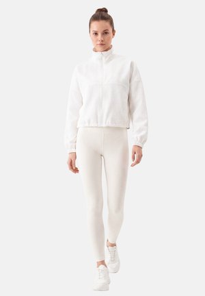 Young woman walking wearing a white zip-up jacket, white leggings, and white sneakers against a plain light background.
