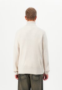 Man wearing cream knitted turtleneck sweater and dark loose jeans, shown from behind against a plain white background.