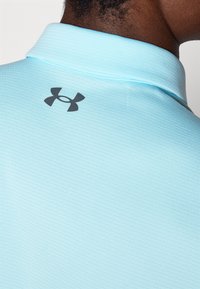 Jasnoniebieska koszulka polo z teksturowanej tkaniny, z czarnym logo Under Armour na plecach kołnierzyka. Klasyczny wzór kołnierzyka.
