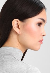 Gullige hoopøreringer med et slankt, forlengt design, hengende fra loben; vist på en modell med langt, mørkt hår og en grå topp.