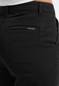 Crne traperice od denima s glatkom teksturom, koje imaju stražnji džep i oznaku marke. Prilagođeni kroj s standardnim šavovima.