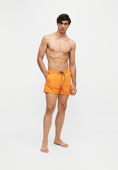 Jeune homme debout pieds nus portant un short de bain orange avec un cordon sur un fond clair uni.