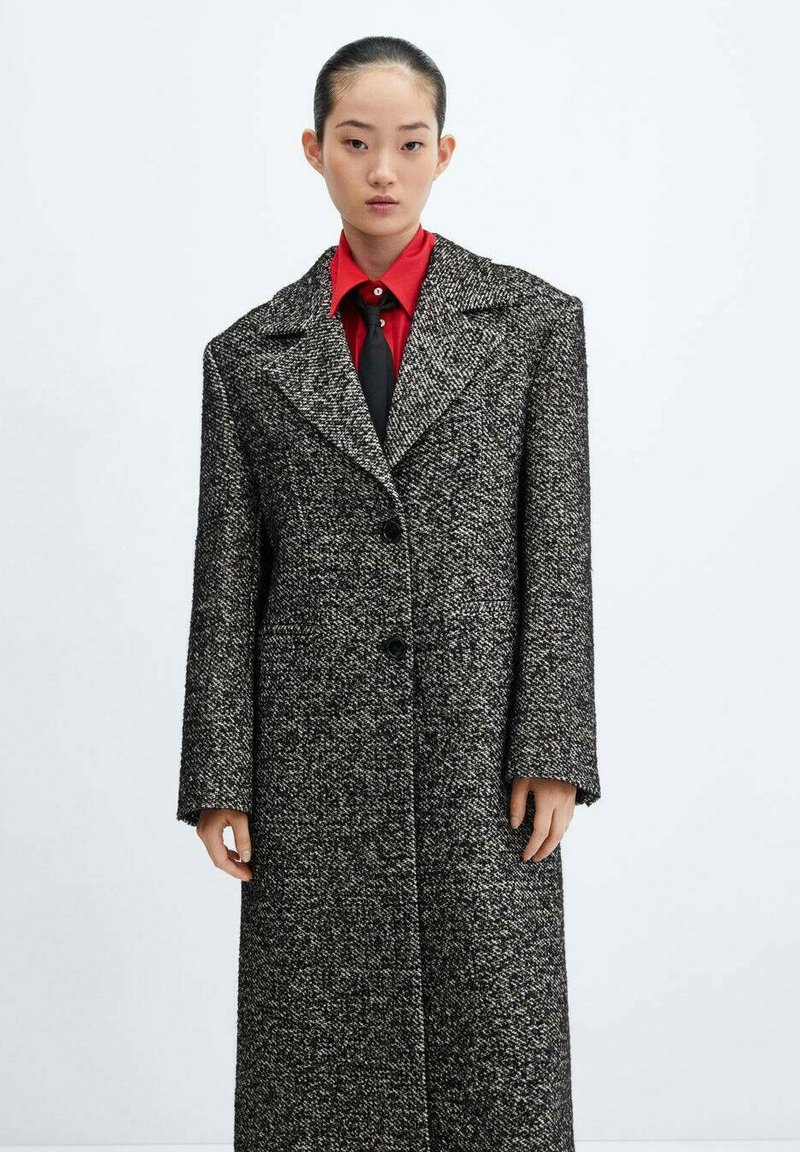 Cappotto lungo in tweed bianco e nero con colletto a punte, bottoni doppi e vestibilità leggermente oversize, indossato sopra una camicia rossa e una cravatta nera.