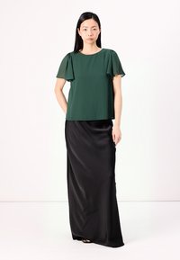 Grüne kurzärmelige Bluse mit durchsichtigen Ärmeln, kombiniert mit einem langen schwarzen Satinrock. Die Bluse hat einen lässigen Schnitt und eine glatte Textur.