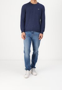 Marinblå crew-neck tröja i mjukt stickat material, parat med ljusblå jeans och vita sneakers. Tröjan har en liten grön logotyp på bröstet.