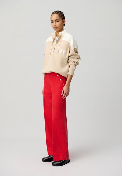 Jersey beige con un collar blanco y logo, combinado con pantalones de pierna ancha rojos de tiro alto con botones. Unos zapatos negros planos completan el atuendo.