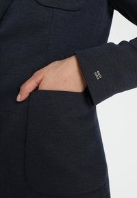 Blazer azul marino con un bolsillo delantero. Los puños tienen un pequeño acento en plata con una "H". La tela parece texturizada y ligeramente elástica.