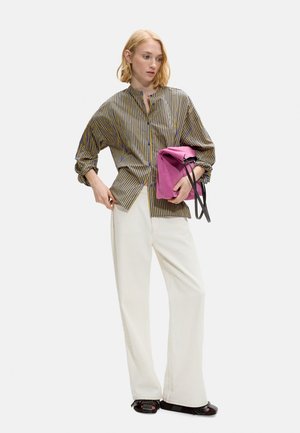 Kobieta w oversize pasekowanej koszuli i szerokich białych spodniach, trzyma złożoną różową kopertówkę przy boku.