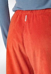Kanskebyxor i livlig orange med ribbad textur, som har ett elastiskt midjeband och en vit etikett med "DEHA" på baksidan.