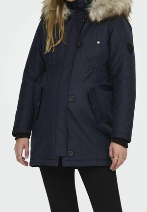 Kvinde iført en mørkeblå vinterjakke med pelsforet hætte, knap- og lynlåslukning samt to forlommer.