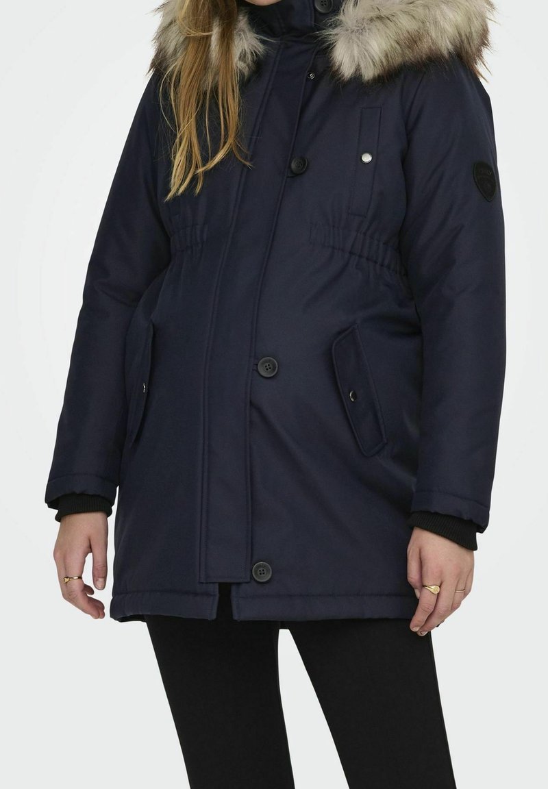 Femme portant un manteau d'hiver bleu marine avec capuche bordée de fourrure, fermeture à boutons et fermeture éclair, et deux poches avant.