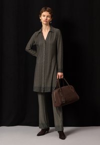 Femme portant une longue chemise à motifs foncés et un pantalon, tenant un sac à main marron sur un fond noir.