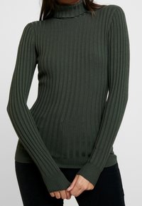 Pull à col roulé côtelé vert foncé avec des manches longues et un design ajusté. Comprend un tissu texturé et des poignets allongés pour une meilleure couverture.