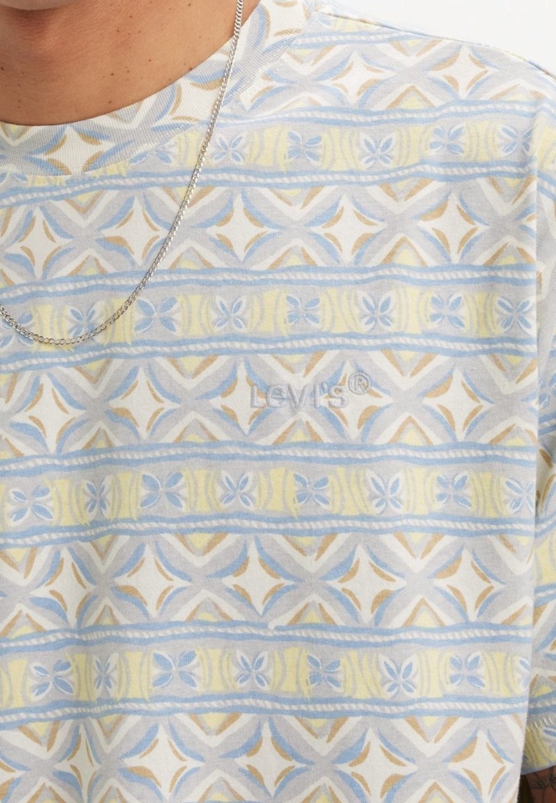 Personne portant une chemise Levi's à motifs avec des formes géométriques bleues, jaunes et beiges et un fin collier en chaîne argentée.