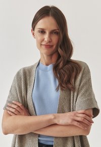 Cardigan en tricot gris sur un haut bleu clair, présentant des manches courtes et une coupe décontractée. Le mannequin pose avec les bras croisés dans un cadre neutre.