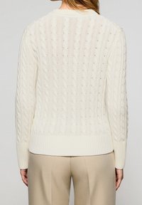 Femme portant un pull en maille torsadée crème et un pantalon beige, vue de dos sur un fond uni.